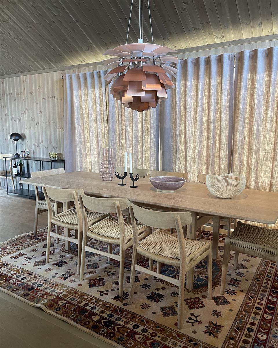 Hytte med lingardiner med stangbånd, lyst trebord og stoler, konglelampe og persisk gulvteppe. Foto.